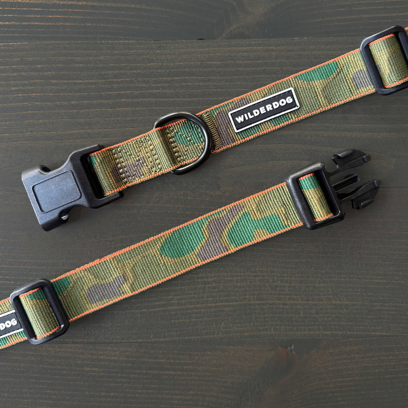 Two "Camo Ultralight Collar" dog collars in green camouflage nylon, featuring black plastic buckles and metal rings with WILDERDOG branding, are displayed on a dark wooden surface.